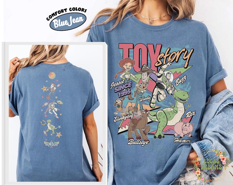 May include: Blue "Comfort Colors" t-shirt featuring a "Toy Story" graphic with characters like Woody, Buzz, and Jessie. The back of the shirt has a vertical design with more characters. The shirt is a light blue color.
