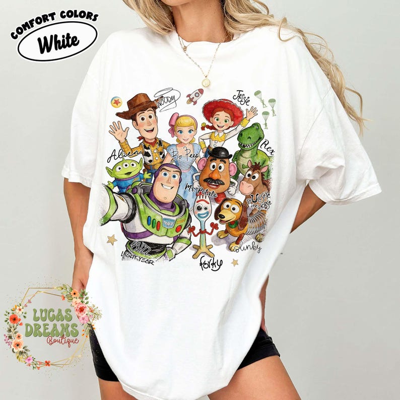 Op de afbeelding: Een wit t-shirt met korte mouwen en een kleurrijke grafische print van Toy Story-personages, waaronder Woody, Buzz Lightyear, Jessie, Bo Peep, Mr. Potato Head, Rex, Bullseye, Slinky Dog, Forky en een Alien, met de namen in cursief schrift.