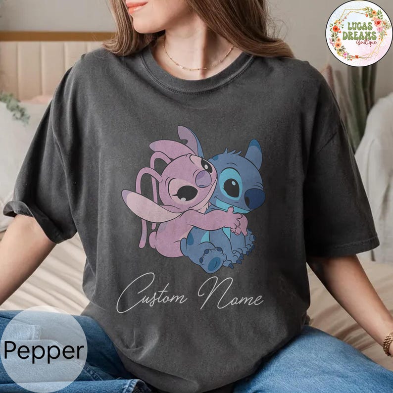 May include: Dark gray t-shirt featuring a cartoon illustration of Stitch and Angel hugging. The design includes the text "Custom Name" in a cursive font. The shirt is a relaxed fit.