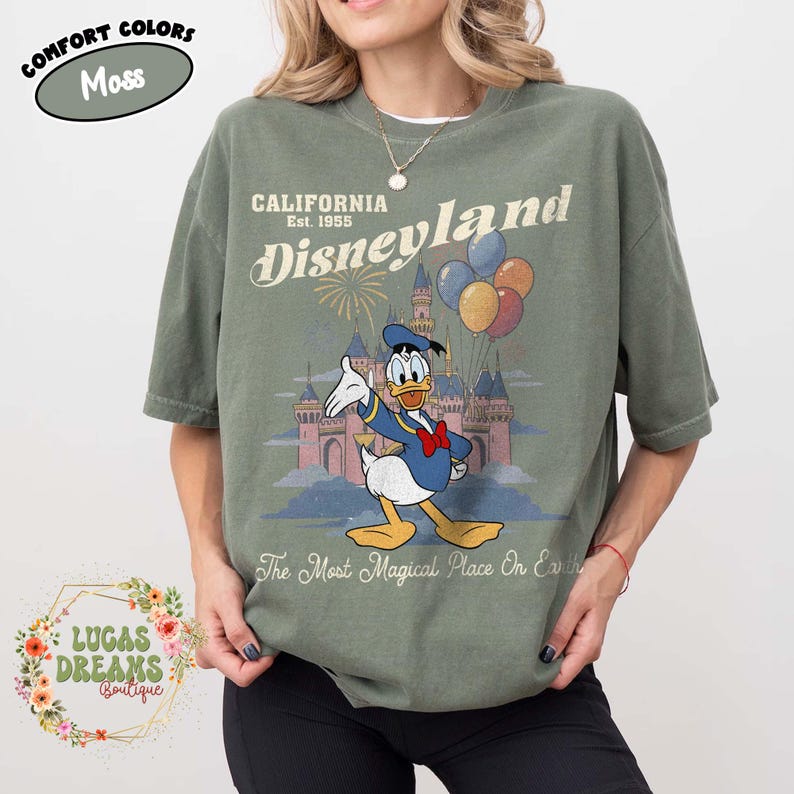 May include: A moss green t-shirt featuring a graphic of Donald Duck in front of a castle with fireworks and balloons. The text "CALIFORNIA Disneyland Est. 1955" and "The Most Magical Place On Earth" are printed on the shirt.