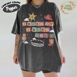 May include: A charcoal gray short-sleeve t-shirt featuring a graphic print of colorful alphabet blocks spelling I KNEW IT I KNEW YOU, a sheriff badge with text INFINITY AND BEYOND, a cowboy hat, a pair of cowboy boots, and a beaded bracelet with text.