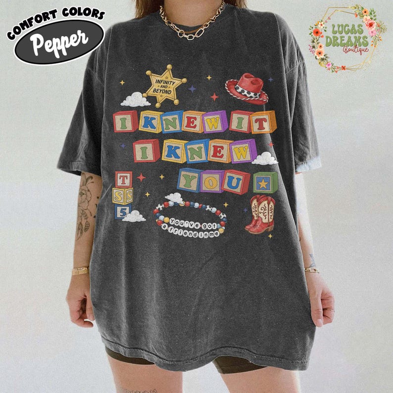 May include: A charcoal gray short-sleeve t-shirt featuring a graphic print of colorful alphabet blocks spelling I KNEW IT I KNEW YOU, a sheriff badge with text INFINITY AND BEYOND, a cowboy hat, a pair of cowboy boots, and a beaded bracelet with text.