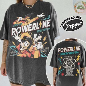 Puede incluir: Camiseta gris oscuro de Powerline World Tour con personajes de dibujos animados y gr&aacute;ficos de rayos. La parte delantera muestra el nombre de la banda y "Stand Out". La parte trasera enumera las ubicaciones de la gira. Hecho con Comfort Colors.
