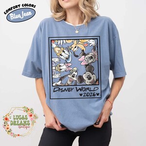 Op de afbeelding: Blauw T-shirt met korte mouwen met een afbeelding van Disney-personages zoals Mickey Mouse, Donald Duck en Goofy. De afbeelding is omlijst met de woorden "DISNEY WORLD 2026". Het shirt heeft de woorden "COMFORT COLORS Blue Jean" linksboven.