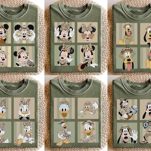 May include: Olive green t-shirts featuring cartoon characters in safari attire. The shirts showcase Mickey Mouse, Minnie Mouse, Pluto, Donald Duck, Daisy Duck, and Goofy in various poses, with safari hats and binoculars.