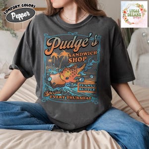 Pu&ograve; includere: T-shirt grigio scuro con grafica per "Pudge's Sandwich Shop". Il design include un pesce dei cartoni animati, palme e le parole "Peanut Butter Special Every Thursday". La maglietta ha un aspetto vintage e usurato.