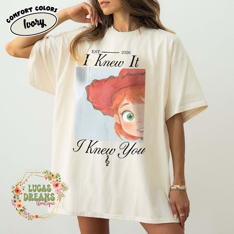 May include: An ivory t-shirt featuring a graphic print of a cartoon character with red hair and a red hat. The text above the graphic reads "EST 2026 I Knew It" and the text below reads "I Knew You" with a small musical note symbol included.