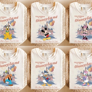 May include: Cream-colored t-shirts featuring Disneyland graphics with various Disney characters. Each shirt displays the Disneyland logo, a castle illustration, and the phrase "The Most Magical Place On Earth."