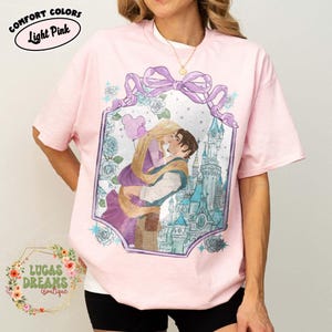 Puede incluir: Camiseta rosa claro con una ilustraci&oacute;n enmarcada de una pareja bes&aacute;ndose frente a un castillo. La ilustraci&oacute;n est&aacute; rodeada de rosas azules y un lazo morado. El texto "COMFORT COLORS Light Pink" est&aacute; en la parte superior.