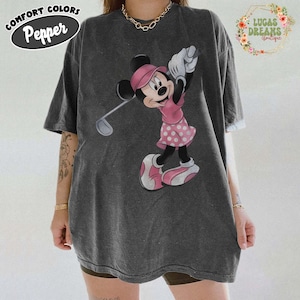 May include: Dark gray t-shirt featuring a Minnie Mouse graphic in pink golfing attire. The shirt has the words "COMFORT COLORS Pepper" printed on the top left. The shirt also has the words "LUCAS DREAMS boutique" printed on the top right.