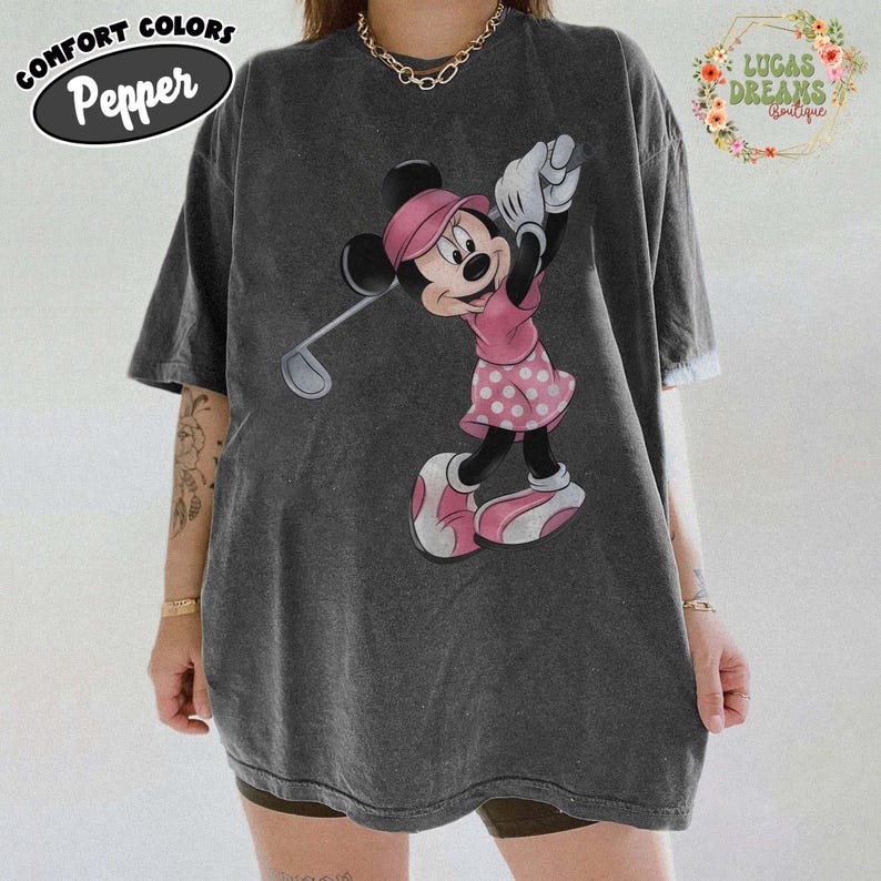 May include: Dark gray t-shirt featuring a Minnie Mouse graphic in pink golfing attire. The shirt has the words "COMFORT COLORS Pepper" printed on the top left. The shirt also has the words "LUCAS DREAMS boutique" printed on the top right.