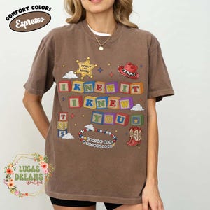 May include: A brown short-sleeve t-shirt featuring a graphic print of colorful alphabet blocks spelling I KNEW IT I KNEW YOU. The design includes a yellow sheriff star with the text INFINITY AND BEYOND, a red cowboy hat, a beaded bracelet with the text YOU'VE GOT A FRIEND IN ME, and a pair of red cowboy boots.