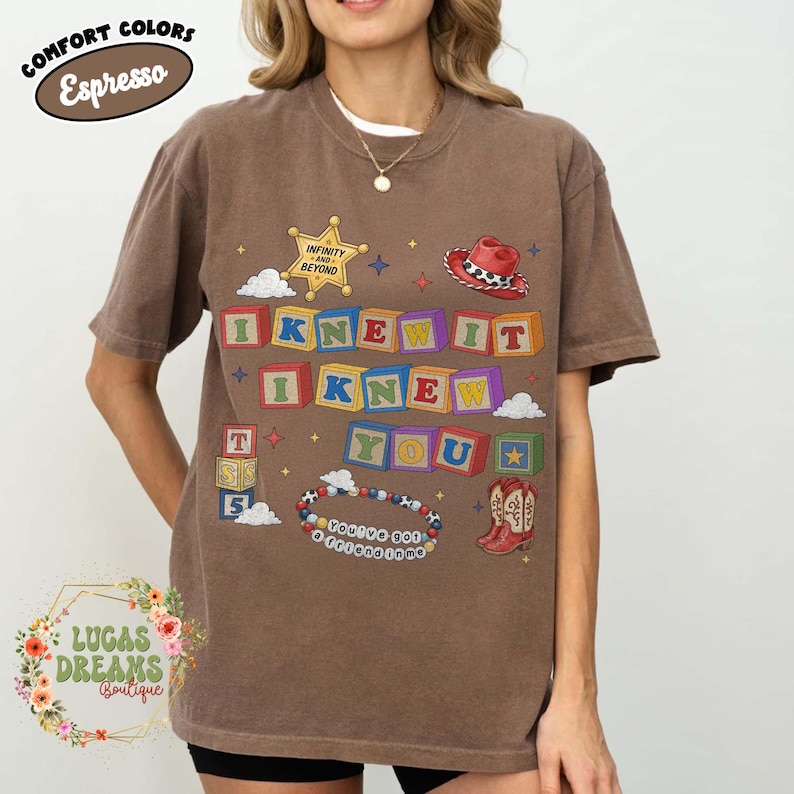 May include: A brown short-sleeve t-shirt featuring a graphic print of colorful alphabet blocks spelling I KNEW IT I KNEW YOU. The design includes a yellow sheriff star with the text INFINITY AND BEYOND, a red cowboy hat, a beaded bracelet with the text YOU'VE GOT A FRIEND IN ME, and a pair of red cowboy boots.