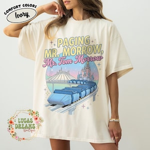May include: Cream-coloured t-shirt featuring the text "Paging Mr. Morrow, Mr. Tom Morrow" above a graphic of a ride and a castle. The shirt has short sleeves and a loose fit. The shirt is from Lucas Dreams Boutique.