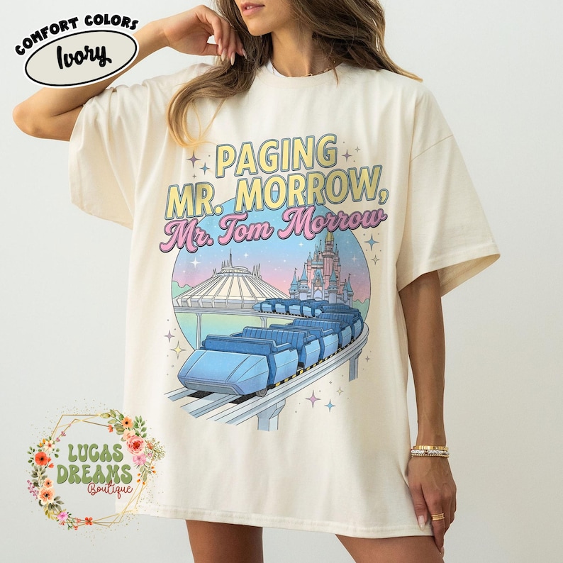 May include: Cream-coloured t-shirt featuring the text "Paging Mr. Morrow, Mr. Tom Morrow" above a graphic of a ride and a castle. The shirt has short sleeves and a loose fit. The shirt is from Lucas Dreams Boutique.