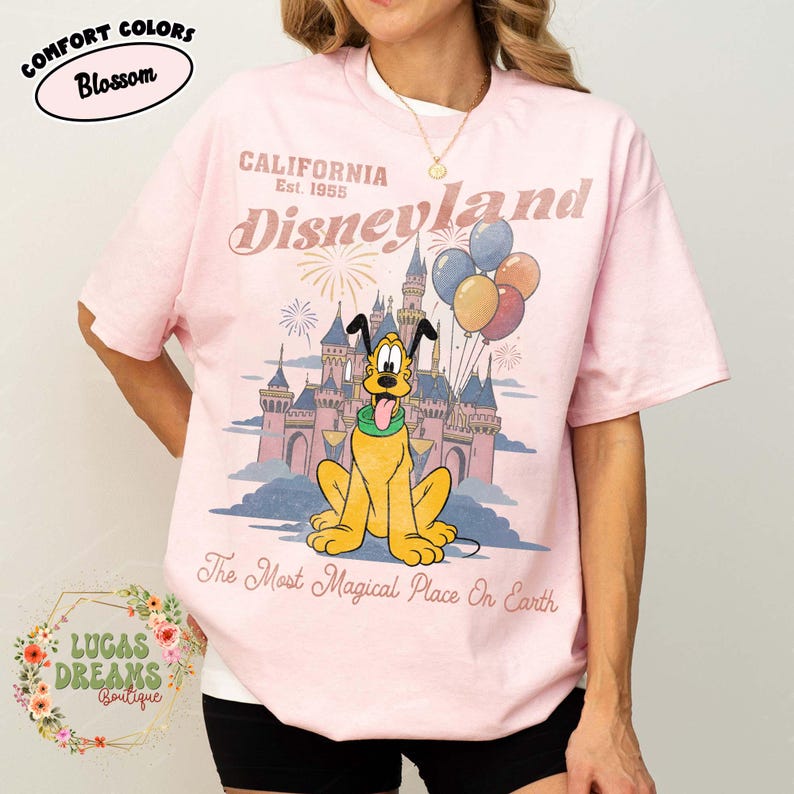 May include: Pink t-shirt with a Disneyland graphic featuring Pluto in front of a castle, balloons, and fireworks. Text includes "California Est. 1955" and "The Most Magical Place On Earth."