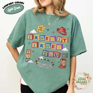 May include: A light green short-sleeve t-shirt featuring a graphic print with colorful alphabet blocks spelling I KNEW IT I KNEW YOU, a sheriff badge with text INFINITY AND BEYOND, a red cowboy hat, a beaded bracelet with text YOU'VE GOT A FRIEND IN ME.