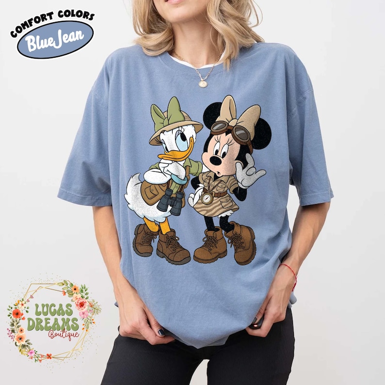 May include: Pale blue t-shirt with Donald Duck and Minnie Mouse in safari attire. The cartoon characters are wearing hats, vests, and boots. The shirt has the text "COMFORT COLORS Blue Jean" at the top and "LUCAS DREAMS Boutique" at the bottom.