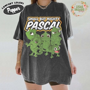 Puede incluir: Camiseta gris oscuro con el texto "SMALL BUT MIGHTY PASCAL" en amarillo y blanco. La camiseta presenta un gráfico de varios camaleones verdes de dibujos animados. La camiseta tiene las palabras "COMFORT COLORS Pepper" en un óvalo negro.