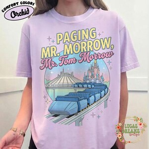 May include: A lilac t-shirt featuring the text "Paging Mr. Morrow, Mr. Tom Morrow" in yellow and pink. The design includes a monorail, Space Mountain, and a castle. The shirt is branded Comfort Colors.