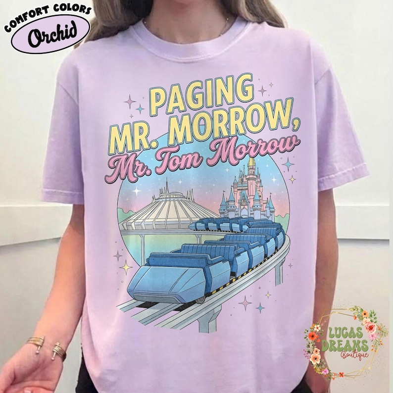 May include: A lilac t-shirt featuring the text "Paging Mr. Morrow, Mr. Tom Morrow" in yellow and pink. The design includes a monorail, Space Mountain, and a castle. The shirt is branded Comfort Colors.