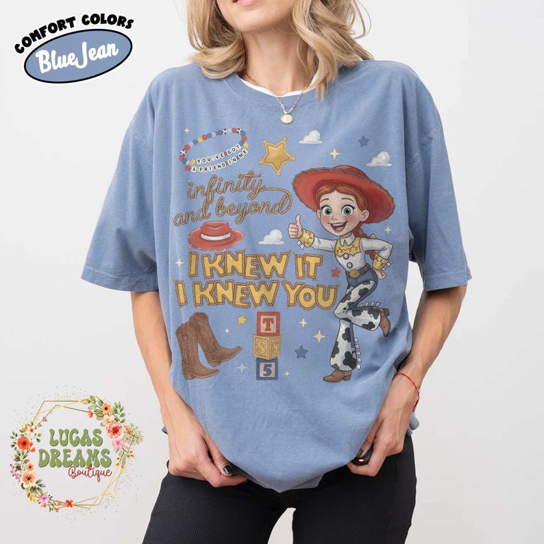 May include: A blue jean colored t-shirt featuring a graphic of the character Jessie from Toy Story. The design includes the text "infinity and beyond", "I KNEW IT I KNEW YOU", a cowboy hat, brown boots, a star, and a stack of blocks with the letters T, S, and 5.