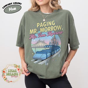 May include: Olive green t-shirt with a graphic featuring a monorail, Space Mountain, and Cinderella's castle. The text reads "Paging Mr. Morrow, Mr. Tom Morrow." The shirt also has the text "Comfort Colors" and "Lucas Dreams Boutique."