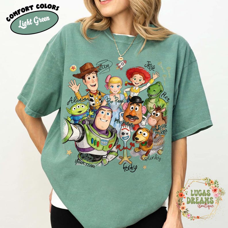 Op de afbeelding: Een lichtgroen T-shirt met korte mouwen en een grafische print van Toy Story-personages, waaronder Woody, Buzz Lightyear, Jessie, Bo Peep, Rex, Mr. Potato Head, Slinky Dog, Bullseye, Forky en een Alien, met de namen in cursief schrift.