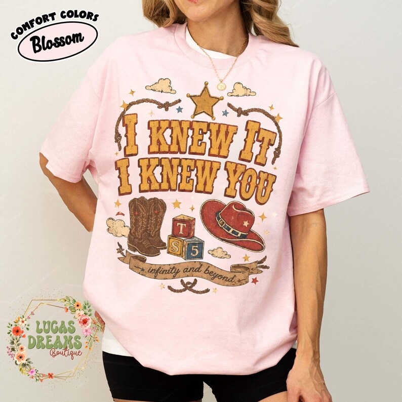 May include: A light pink t-shirt featuring a graphic print with the text I KNEW IT I KNEW YOU and infinity and beyond. The design includes a brown cowboy boot, a red cowboy hat, a star, and two alphabet blocks labelled T, S, and 5 in a rustic style.