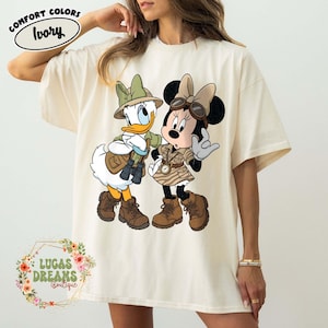 May include: Ivory Comfort Colors t-shirt featuring Donald Duck and Minnie Mouse in safari outfits. Donald Duck wears a green hat and khaki jacket, while Minnie Mouse sports a safari dress. The shirt has the text "LUCAS DREAMS BOUTIQUE".