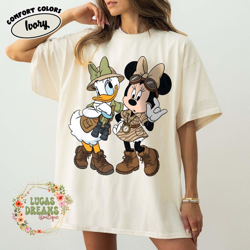 May include: Ivory Comfort Colors t-shirt featuring Donald Duck and Minnie Mouse in safari outfits. Donald Duck wears a green hat and khaki jacket, while Minnie Mouse sports a safari dress. The shirt has the text "LUCAS DREAMS BOUTIQUE".