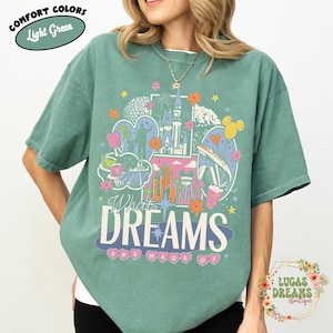 K&ouml;nnte beinhalten: Hellgr&uuml;nes T-Shirt mit einer farbenfrohen Grafik mit einem Schloss, Spaceship Earth und anderen Themenparkelementen. Der Text "What Dreams Are Made Of" ist in einer verspielten Schriftart dargestellt. Das Shirt tr&auml;gt die Aufschrift "Comfort Colors" und "Light Green".