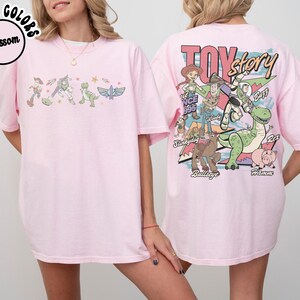 May include: Pale pink t-shirt with a Toy Story theme. The front has a line of characters, and the back features a larger graphic with the film's title and characters. The shirt has a loose fit.