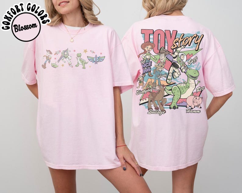 May include: Pale pink t-shirt with a Toy Story theme. The front has a line of characters, and the back features a larger graphic with the film's title and characters. The shirt has a loose fit.