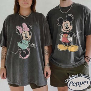 May include: Two charcoal gray oversized t-shirts featuring vintage-style graphic prints. The left shirt displays Minnie Mouse with a pink bow and teal dress, while the right shirt features Mickey Mouse in red shorts and yellow shoes with text.