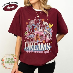 K&ouml;nnte beinhalten: Burgunderfarbenes T-Shirt mit einer farbenfrohen Grafik eines Schlosses, Wahrzeichen und dem Text "What Dreams Are Made Of". Das Shirt hat kurze &Auml;rmel und ein wei&szlig;es Unterhemd. Die Worte "Comfort Colors" und "Maroon" stehen oben in einem Kreis.