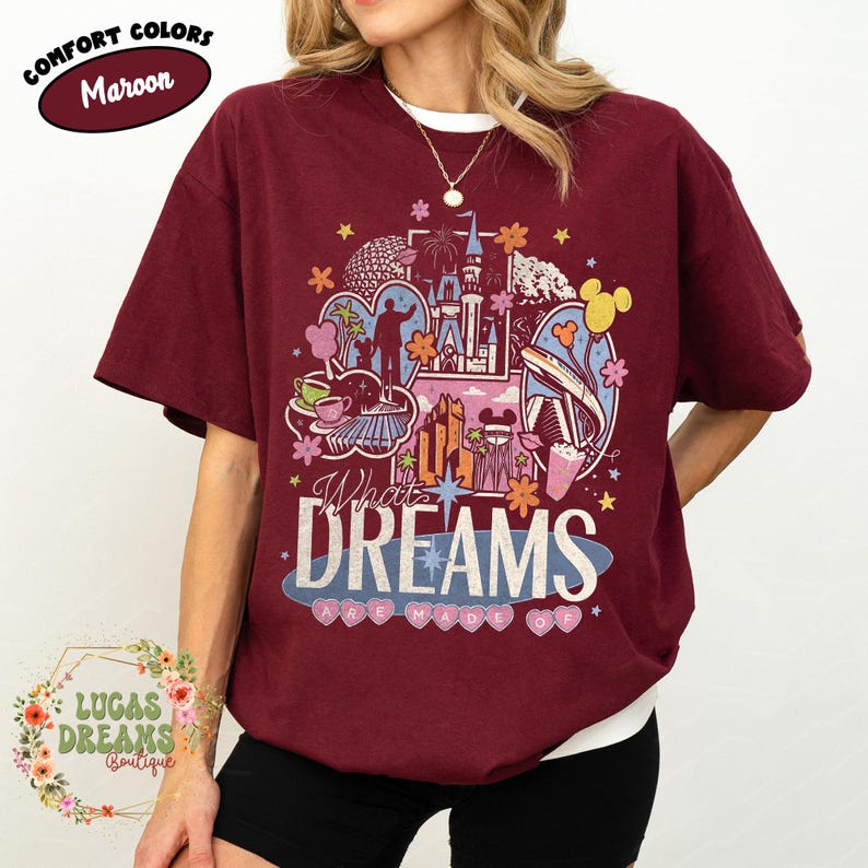 K&ouml;nnte beinhalten: Burgunderfarbenes T-Shirt mit einer farbenfrohen Grafik eines Schlosses, Wahrzeichen und dem Text "What Dreams Are Made Of". Das Shirt hat kurze &Auml;rmel und ein wei&szlig;es Unterhemd. Die Worte "Comfort Colors" und "Maroon" stehen oben in einem Kreis.