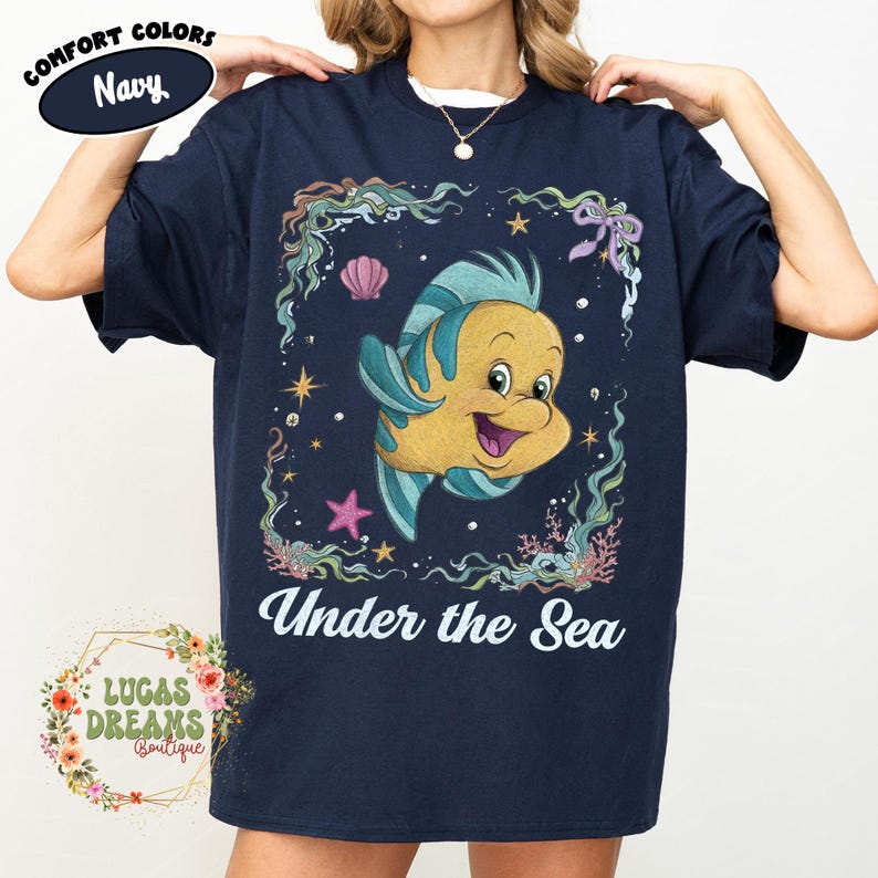 May include: Navy blue t-shirt featuring a cartoon fish with blue and yellow coloring, surrounded by an underwater scene with the text "Under the Sea". The shirt also has the text "Comfort Colors" and "Lucas Dreams Boutique".