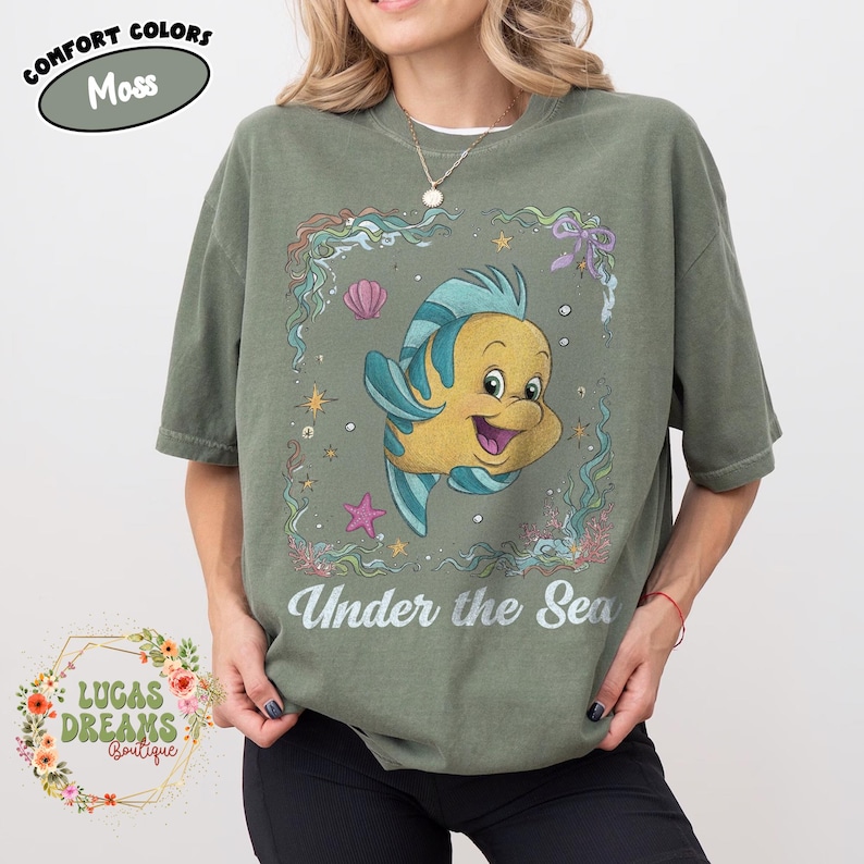 May include: Moss green t-shirt featuring a cartoon image of a yellow and blue fish inside a decorative frame with the words "Under the Sea". The shirt also has the words "Comfort Colors" and "Lucas Dreams Boutique".