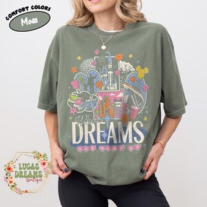 K&ouml;nnte beinhalten: Moosgr&uuml;nes T-Shirt mit einem farbenfrohen Grafikdesign mit einem Schloss, Wahrzeichen und dem Text "What Dreams Are Made Of". Das Shirt hat kurze &Auml;rmel und einen Rundhalsausschnitt. Die Worte "Comfort Colors" und "Moss" sind in einem Kreis oben.