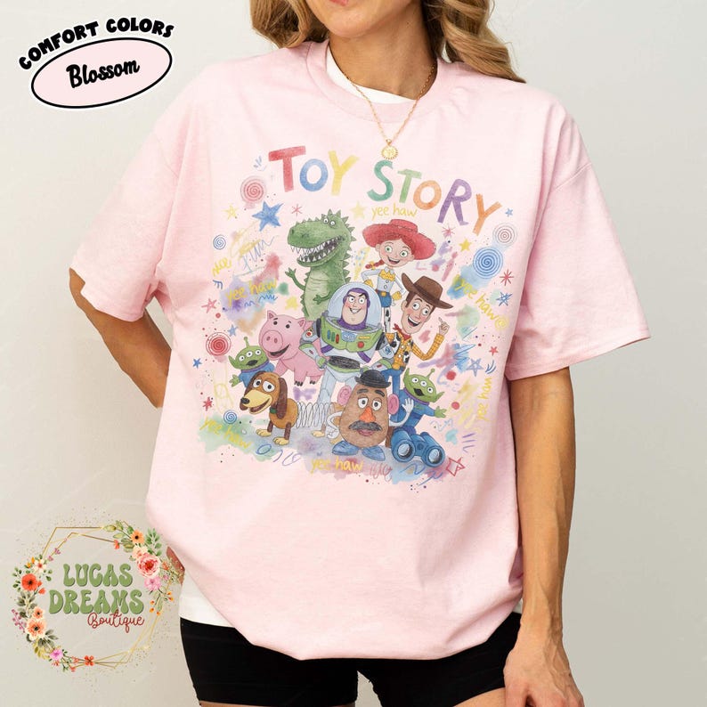Pu&ograve; includere: Una maglietta rosa chiaro con la scritta "Toy Story" in lettere colorate e una grafica dei personaggi del film d'animazione. La maglietta ha maniche corte e girocollo. Il testo "Comfort Colors Blossom" &egrave; in un ovale rosa.