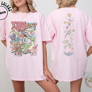 May include: A light pink t-shirt featuring a colorful graphic of Toy Story characters. The front design includes Woody, Buzz Lightyear, and other characters with the text "Toy Story" and "Since 1995". The back of the shirt has additional character graphics.
