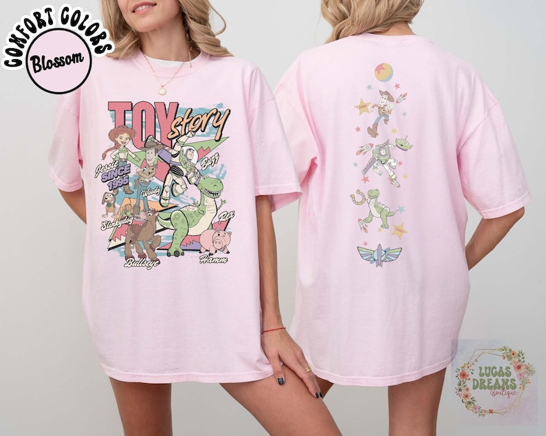 May include: A light pink t-shirt featuring a colorful graphic of Toy Story characters. The front design includes Woody, Buzz Lightyear, and other characters with the text "Toy Story" and "Since 1995". The back of the shirt has additional character graphics.