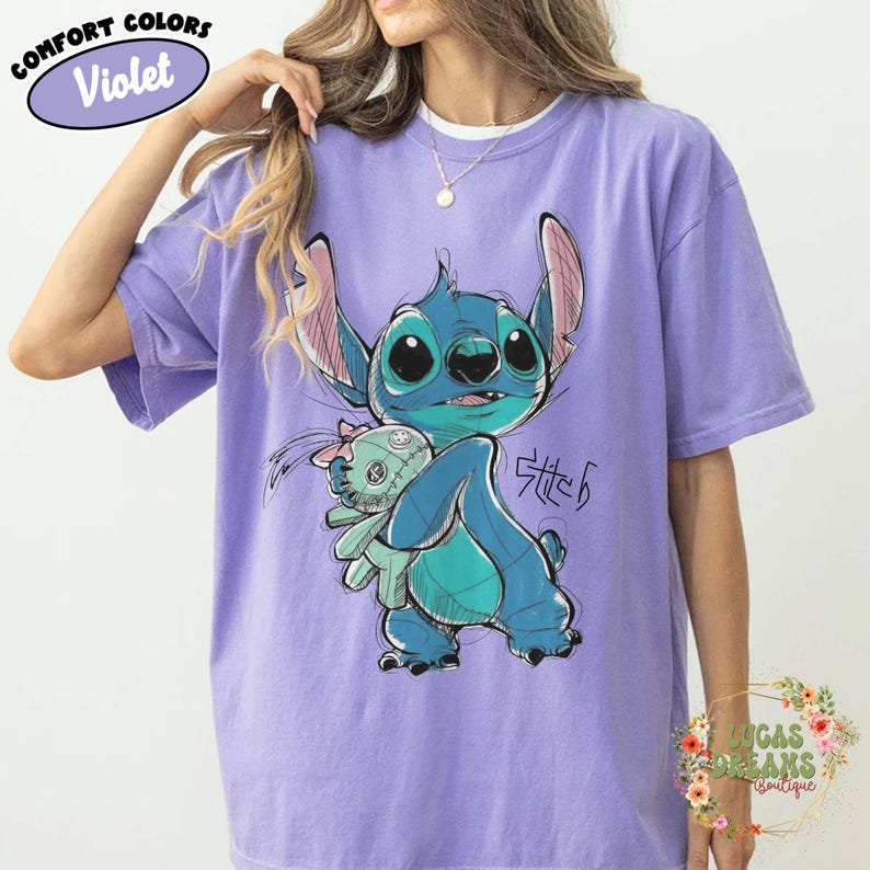 May include: A violet short-sleeve t-shirt featuring a graphic illustration of the blue character Stitch holding a small green doll. The design includes a sketch-like aesthetic with dark blue outlines and the handwritten text Stitch in black ink.