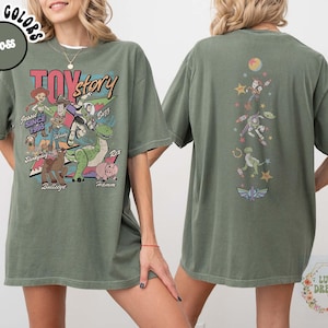 May include: Moss green t-shirt featuring a graphic of Toy Story characters, including Woody, Buzz Lightyear, and Rex. The front design includes the text "Toy Story" and character names. The back of the shirt has a vertical design with more characters and a logo.