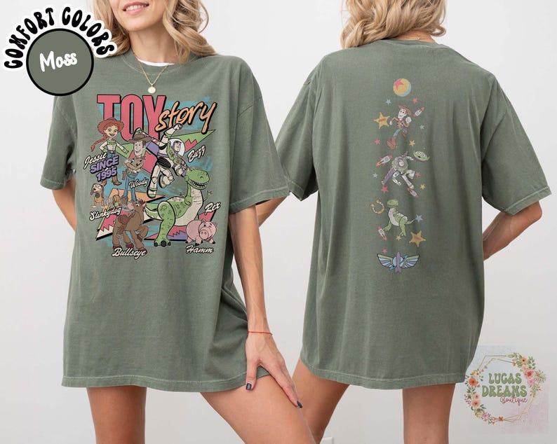 May include: Moss green t-shirt featuring a graphic of Toy Story characters, including Woody, Buzz Lightyear, and Rex. The front design includes the text "Toy Story" and character names. The back of the shirt has a vertical design with more characters and a logo.