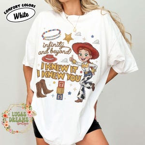 May include: A white t-shirt featuring a graphic print of a cowgirl character, a red hat, brown cowboy boots, alphabet blocks, and a beaded bracelet. The text reads infinity and beyond and I KNEW IT I KNEW YOU. The shirt has short sleeves and a crew neck.