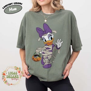 May include: Moss green t-shirt featuring a cartoon Daisy Duck dressed as a mummy for Halloween. Daisy wears a purple bow, bandages, and carries a pumpkin basket. The shirt has the text "COMFORT COLORS" and "Moss".