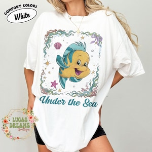 May include: White t-shirt with a cartoon illustration of a yellow and blue fish inside a decorative frame. The text "Under the Sea" is below the fish. The shirt also has the text "Comfort Colors White".
