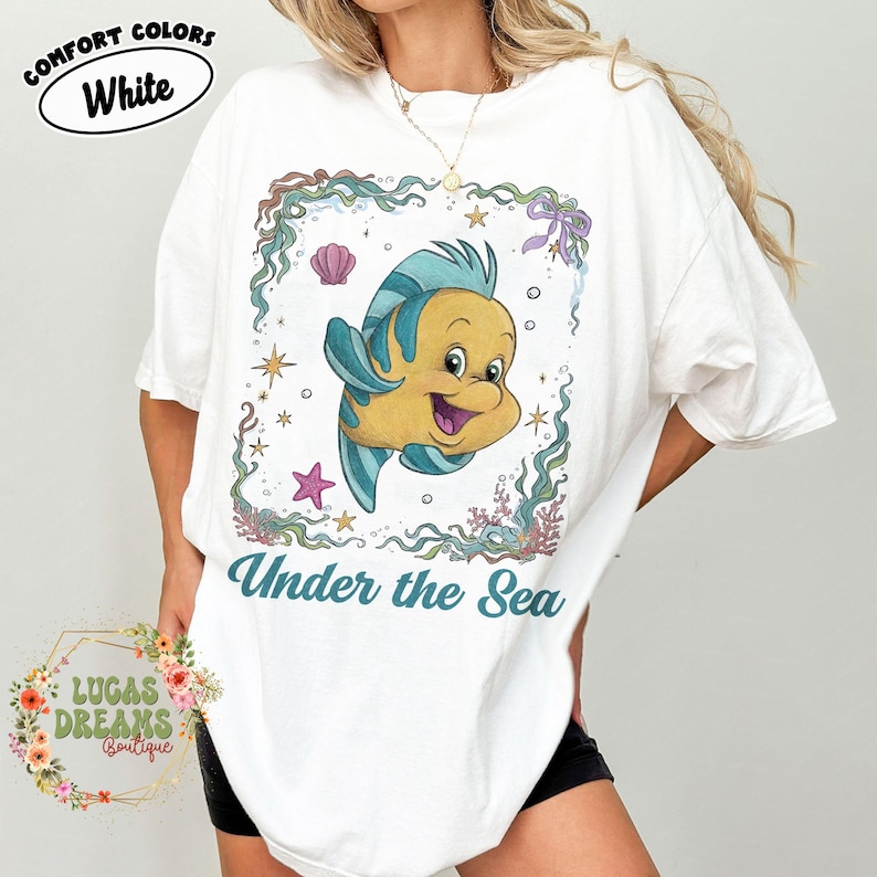 May include: White t-shirt with a cartoon illustration of a yellow and blue fish inside a decorative frame. The text "Under the Sea" is below the fish. The shirt also has the text "Comfort Colors White".